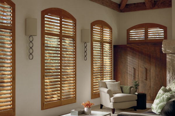 Elegant sitting area with custom arched-top hardwood shutters for light control, textured walls, and neutral furniture in a light-filled architectural space